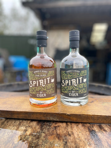 A pair of bottles of spirit say on top of a wooden plank. The one on the left is honey coloured and the label reads Oak Aged Bignose and Beardy Spirit of Cider while the other is clear and the label reads Bignose and Beardy Spirit of Cider.