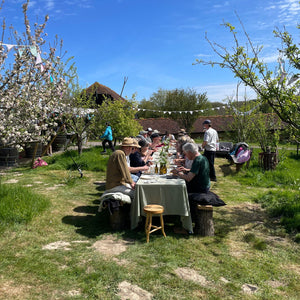 Group of people dining outdoors in a garden setting with trees and clear sky.