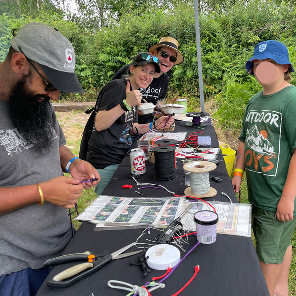 A group of people gathered outdoors around a table set up under a canopy, working on what appears to be a craft or fishing-related project with cords, tools, and printed materials. One person wears a green graphic T-shirt and blue cap, while others are seated at the table handling wires and equipment.
