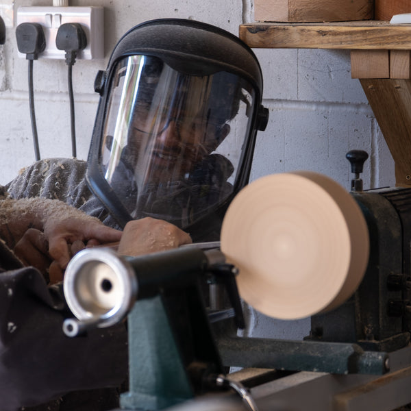 Person using a wood lathe with a protective face shield in a workshop setting.