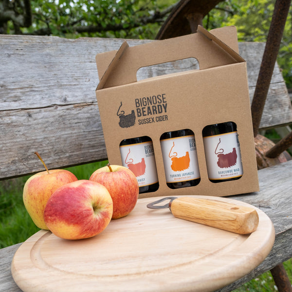 Three beer bottles in a cardboard carrier with apples and a wooden mallet on a rustic wooden table outdoors.