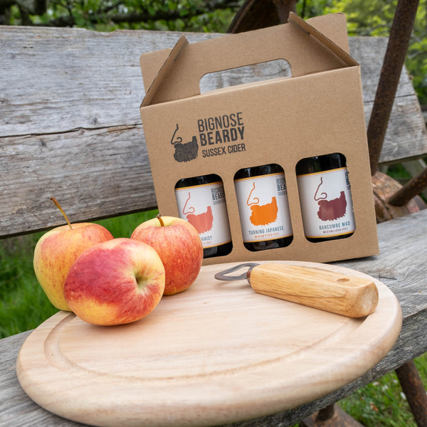 Three beer bottles in a cardboard carrier with apples and a wooden mallet on a rustic wooden table outdoors.