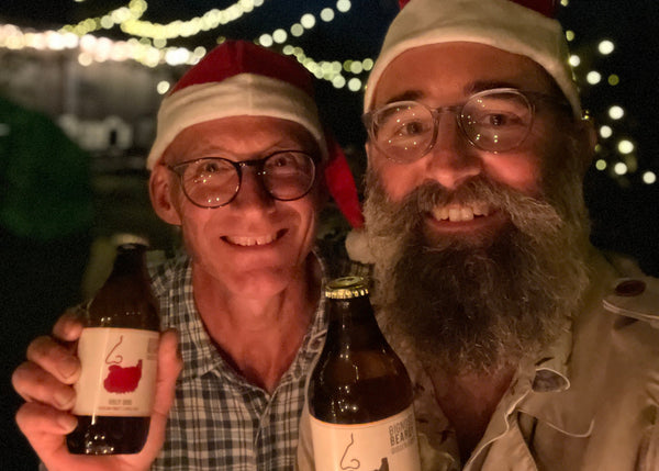 Bignose and Beardy wearing Santa hats holding bottles of cider with festive lights in the background