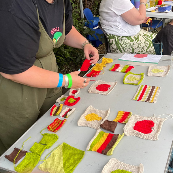 A woman at an outdoor craft table works on handmade knitted items, with other people seated nearby and colorful fabric pieces arranged on the table.