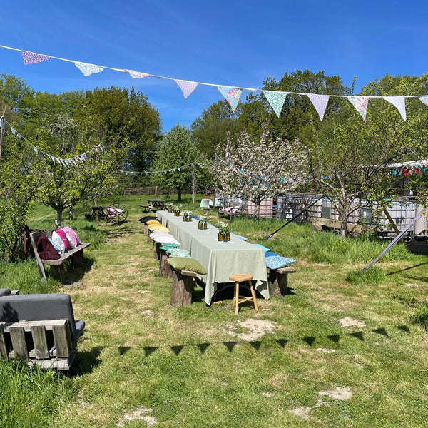 Outdoor setting with a long table covered by a green cloth, surrounded by chairs and bunting flags in a garden.