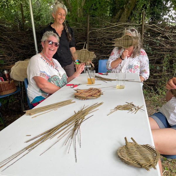 Several people gathered around a white folding table outdoors, working on woven basket or paddle-like crafts made from natural fibers. The scene is set under a canopy with trees and branches in the background.
